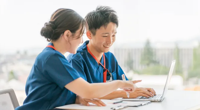 A young woman and man sitting next to each other, wearing scrubs, and studying in front of some papers and a laptop